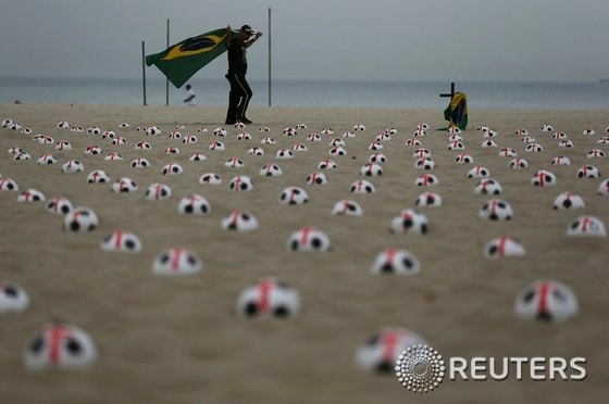 브라질 리우데자네이루의 코파카바나 해변에서 22일(현지시간) 브라질 국기를 든 한 시위자가 빨간 십자가가 그려진 축구공들 사이를 걸어가고 있다. 이 축구공은 '리우 평화'라는 NGO에서 마련한 것인데, 교육, 의료, 공공서비스를 월드컵 경기장과 맞먹는 수준으로 해 줄 것을 정부에 요구하는 의미를 담고 있다.© News1 