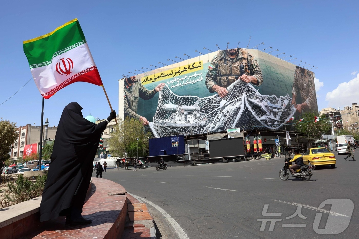 5일(현지시간) 이란 수도 테헤란 엔켈랍 광장에서 한 여성이 "호르무즈 해협은 여전히 ​​폐쇄되어 있다＂라는 문구가 적힌 광고판 옆에 서서 이란 국기를 들고 있다. 2026.04.05. ⓒ AFP=뉴스1